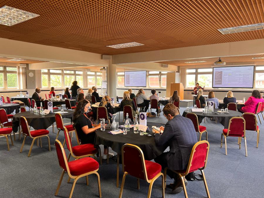 A conference room with a projector displaying content, and people sitting around a rounded table, listening attentively. The table is set with glasses and a glass water bottle, surrounded by red chairs.